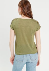 Top verde oliva a maniche corte con dettagli in pizzo sulle spalle, tessuto leggero, vestibilità rilassata, abbinato a jeans a vita alta azzurri.