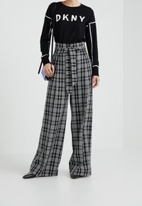 Schwarzer DKNY-Pullover mit weißem Logo, kombiniert mit hochtaillierten karierten Hosen in Schwarz und Weiß, die mit einem Bindegürtel und weitem Bein gestaltet sind.