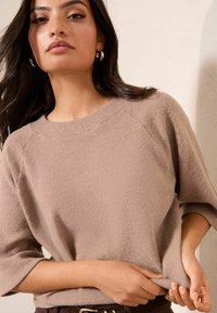 Bežinė, tekstūruota megztinė su apvaliu iškirpimu ir trimis ketvirtadaliais rankovių, derinama su rudomis diržais. Lygus audinys su raižytomis detalėmis.