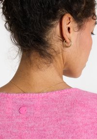 Rosa gestrickter Pullover mit weicher Textur, runder Halslinie und einem kleinen rosa Smiley-Knopfdetail auf der Rückseite. Eine goldene Kettenhalskette ist sichtbar.