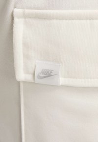 Biały materiałowy haft o teksturowanej powierzchni, z małą białą metką z szarym logo Nike. Widoczny projekt kieszeni.
