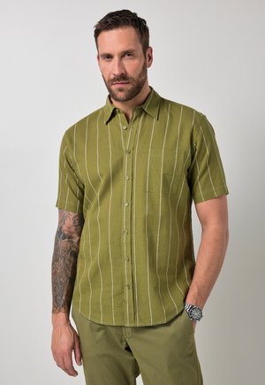 Uomo con barba e braccio sinistro tatuato che indossa una camicia verde a maniche corte con righe bianche e pantaloni abbinati, mano in tasca.