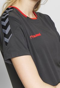 Camiseta deportiva gris de manga corta, con acentos rojos y un patrón de chevrones en negro y blanco en el hombro. Presenta un logo en rojo.