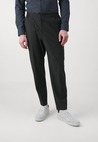 Svarte tapered bukser med knappelukking og elastisk midjebånd, parret med hvite sneakers. Tøyet er glatt med en subtil glans.