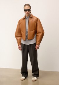 Brūna ādas polsterota jaka ar strukturētām plecu daļām, pelēka svītraina krekla ar garām piedurknēm, tumšas bikses un baltas kurpes ar beigu akcentiem.