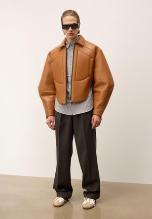 Homme portant une grande veste en cuir marron, une chemise grise rayée, un large pantalon foncé, des baskets beiges et des lunettes de soleil noires, se tenant contre un arrière-plan neutre.