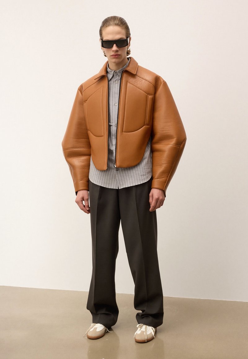 Brūna ādas polsterota jaka ar strukturētām plecu daļām, pelēka svītraina krekla ar garām piedurknēm, tumšas bikses un baltas kurpes ar beigu akcentiem.