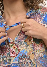 Blusa colorata con motivo a fantasia e scollatura a V profonda, caratterizzata da intrecci intricati in blu, rosso e verde. Tessuto leggero con lacci.