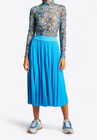 Långärmad topp med blommönster och hög hals, kombinerad med en plisserad, ljusblå midikjol och vita sneakers med färgglada detaljer.