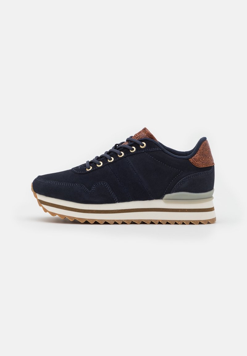 Baskets en daim bleu marine avec des accents en cuir marron au talon et à la languette, semelle rayée blanche et beige, et lacets noirs avec œillets dorés.