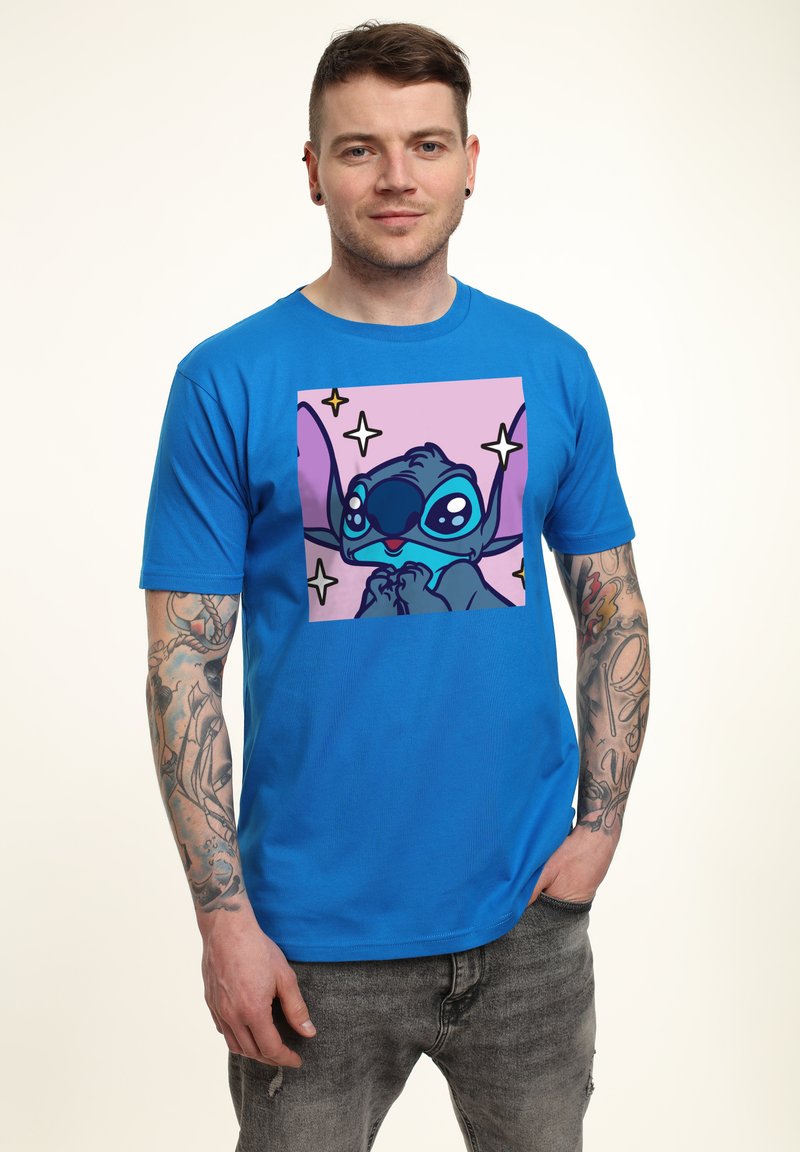 Mann mit tätowierten Armen trägt ein blaues T-Shirt mit dem Cartoon-Charakter Stitch vor einem pinken quadratischen Hintergrund mit Sternen.