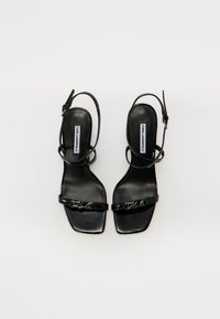 Sandales en cuir verni noir avec des bouts carrés, des lanières fines à la cheville et un logo accentuant la lanière avant. Texture lisse et brillante.