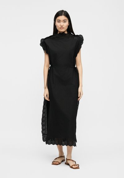 Γυναίκα που στέκεται, φοράει μαύρο αμάνικο midi φόρεμα με διακοσμητική δαντέλα με τρύπες, πλαινούς δεσμούς και καφέ επίπεδα σανδάλια, σε απλό φόντο.