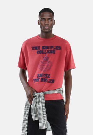 Rød bomulds t-shirt med marineblå tekst "THE KOOPLES COLLEGE" og "ABOVE THE RULES," båret med en sort og hvidternet skjorte knyttet om livet.