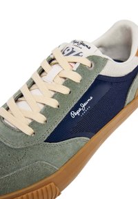 Zelená semišová teniska s tmavomodrými sieťovinovými panelmi, béžovými šnúrkami a gumovou podrážkou. Na boku je značka a textúrované detaily.