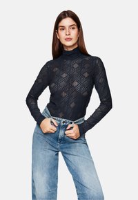 Schwarzes Spitzenpoloshirt mit geometrischen Mustern; taillierte lange Ärmel; kombiniert mit hoch geschnittenen hellblauen Jeans.