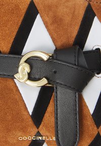 Geantă din suede maro cu un design geometric alb-negru, având o curea din piele neagră cu o cataramă aurie și logo-ul brandului.