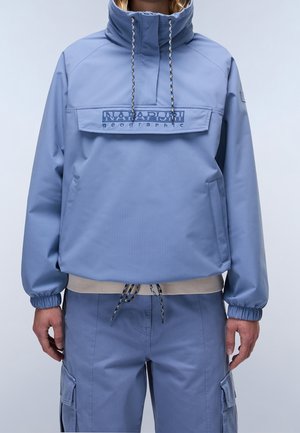 Person trägt eine hellblaue Napapijri Pullover-Jacke mit Kordelzugkapuze und passende Cargohosen, steht vor einem schlichten Hintergrund.