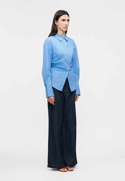 Nő áll előre nézve, kék csíkos gombos ingben, nagy mandzsettákkal, sötét színű, széles szárú nadrágban és szandálban.