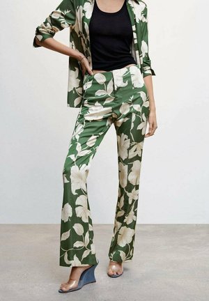 Frau trägt grüne, geblümte Hose mit weitem Bein und passenden Blazer über einem schwarzen Tanktop, kombiniert mit transparenten Keilsandalen.