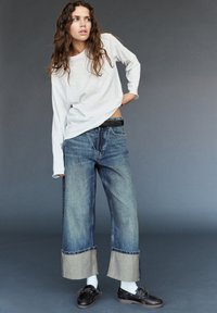 Ženska z dolgimi kodrastimi lasmi, oblečena v belo dolg rokav, široke zvite kavbojke, bele nogavice in črne čolnaste čevlje, stoji ob sivi ozadju.