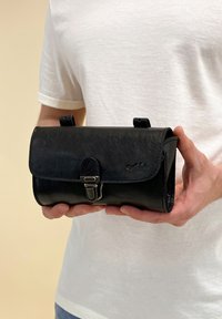 Sort lædertaske med flap-lukning og metalspænde. Har to løkker på toppen. Glat tekstur med subtil korn-detail.