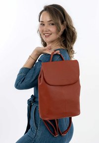 Roter Leder-Rucksack mit strukturiertem Finish, Klappenverschluss und verstellbaren Trägern, gehalten von einer Person im Denim-Outfit.