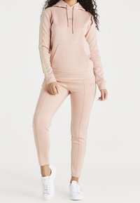 Lichtroze hoodie met gouden strepen op de mouwen, voorzak, gecombineerd met bijpassende taps toelopende broek. Witte sneakers maken de outfit compleet.