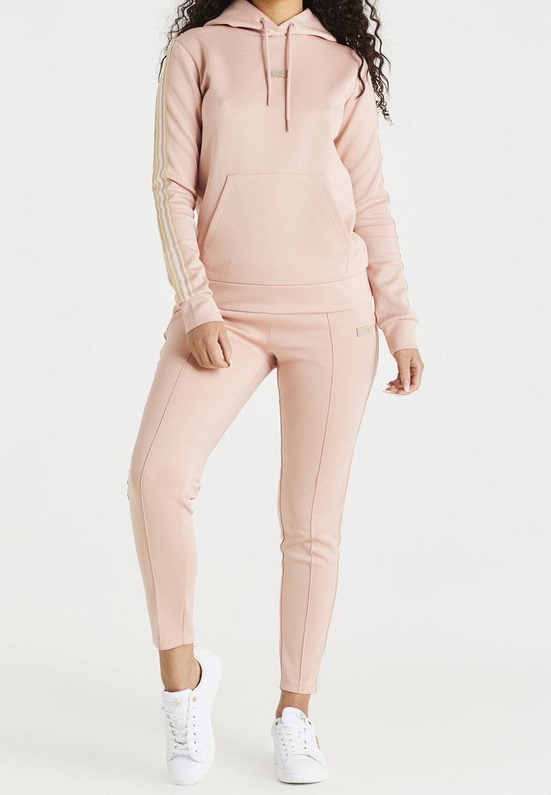 Jasnoróżowa bluza z kapturem ze złotymi paskami na rękawach, przednia kieszeń, w połączeniu z dopasowanymi zwężanymi spodniami. Całość dopełniają białe trampki.