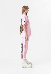 Camiseta rosa de gran tamaño con un colorido diseño bordado, combinada con pantalones de chándal rosas que presentan patrones de alas y una etiqueta lateral. Botas blancas de suela gruesa.