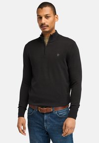 Sort zip-hals sweater lavet af glat stof, med en tætsiddende design, ribbede manchetter og et lille logo på brystet. Båret med blå jeans.