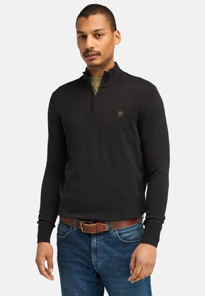 Black zip-neck sweater made of smooth fabric, featuring a fitted design, ribbed cuffs, and a small logo on the chest. Worn with blue jeans.