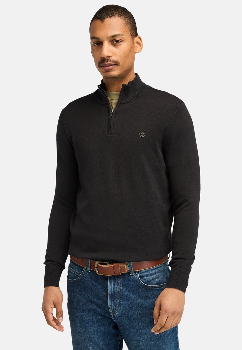 Sort zip-hals sweater lavet af glat stof, med en tætsiddende design, ribbede manchetter og et lille logo på brystet. Båret med blå jeans.