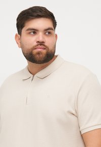Beige polo shirt with a texture pattern, short sleeves, and a front zipper. Features a small logo on the chest.