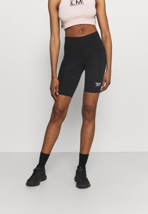 Kobieta ubrana w czarne krótkie spodenki rowerowe Reebok, czarne skarpetki, czarne trampki oraz jasnoróżowy krótki top na ramiączkach z logo "LM".