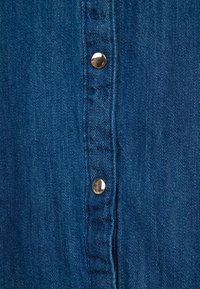 Tessuto di denim blu scuro con una fila di bottoni metallici argentati, che mostra una texture liscia e cuciture visibili lungo la pattina.