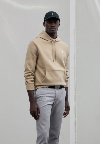 Beige hoodie met capuchon en buidelzak, voorzien van trekkoorden, gecombineerd met lichtgrijze broek. Zwarte pet met een geel logo.