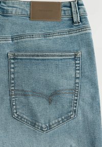 Lyseblå denimjeans med en brun læderlap med mærket "DENIM SUPPLY" og en baglomme med kontrasterende syninger i et buet mønster.