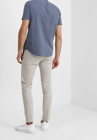 Camicia a maniche corte, con bottoni, in tessuto blu scuro abbinata a pantaloni slim-fit beige chiaro e sneakers bianche, vista da dietro.