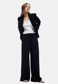 Zwarte blazer op maat over een witte tanktop, gecombineerd met losse zwarte broeken van een geweven stof en een elastische tailleband. Zilveren sandalen.