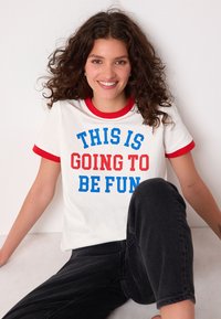 Maglietta bianca con scollo e maniche a contrasto rosse, con testo blu e rosso che recita "QUESTO SARÀ DIVERTENTE." Vestibilità casual.