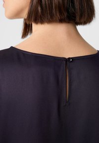 Blusa blu navy con scollatura tonda e un piccolo apertura sul retro chiusa da un bottone scuro, realizzata in un tessuto liscio e strutturato.