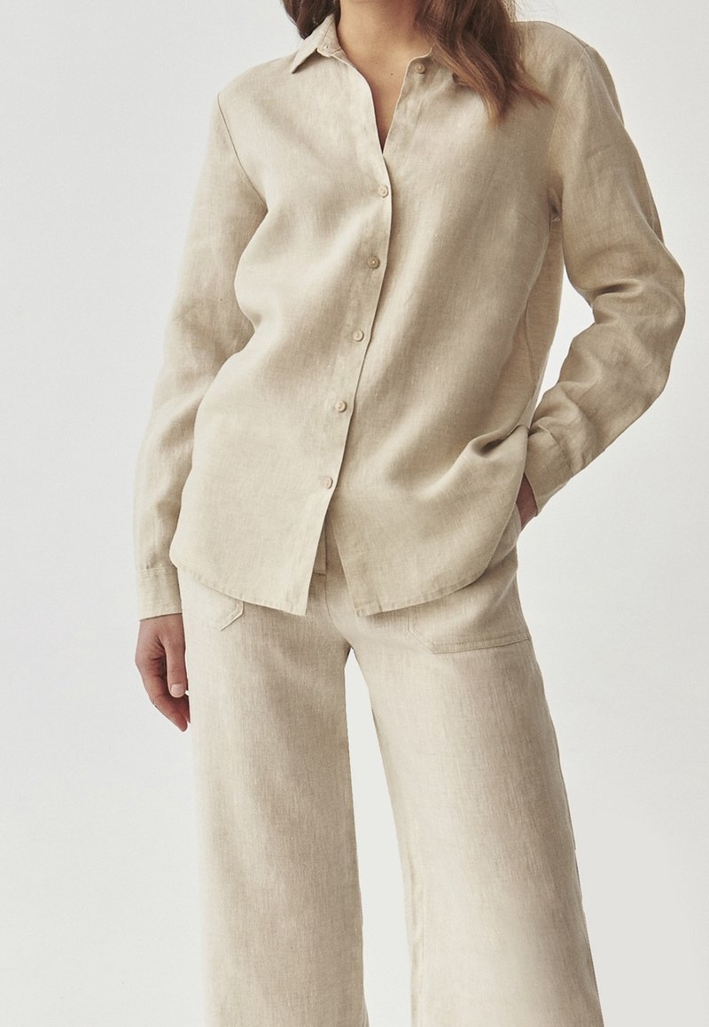 Femme portant une chemise en lin beige à boutons et un pantalon à jambes larges assorti, debout avec une main dans la poche devant un fond blanc.