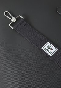 Borsa nera con texture, dotata di una tracolla in nylon e di un gancio metallico. Presenta un'etichetta con il logo Lacoste ricamato, con un design di coccodrillo in verde e rosso.