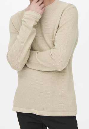 Homme en pull beige en maille à manches longues, un bras croisé et l'autre main touchant le menton, portant un pantalon noir sur fond blanc.