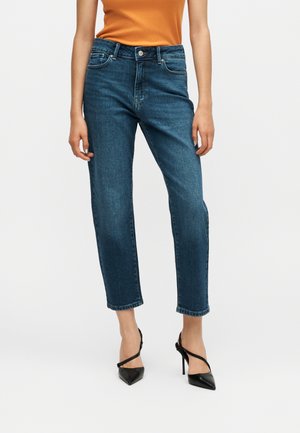 Donna che indossa jeans blu a gamba dritta, un top arancione senza maniche e scarpe nere con tacco alto, in piedi contro uno sfondo semplice.