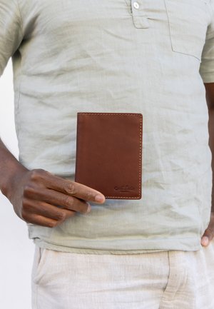 Person in hellem Hemd und Hose hält eine geschlossene braune Lederbrieftasche mit sichtbaren Nähten und geprägtem Logo.