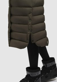 Puffer coat in dark olive green with horizontal stitching, snap closures, and side slits, paired with black Moon Boots featuring a glossy finish.