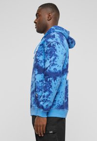 Sweat à capuche bleu tie-dye avec une poche kangourou, capuche réglable à cordon, poignets côtelés et un logo blanc audacieux sur le devant.