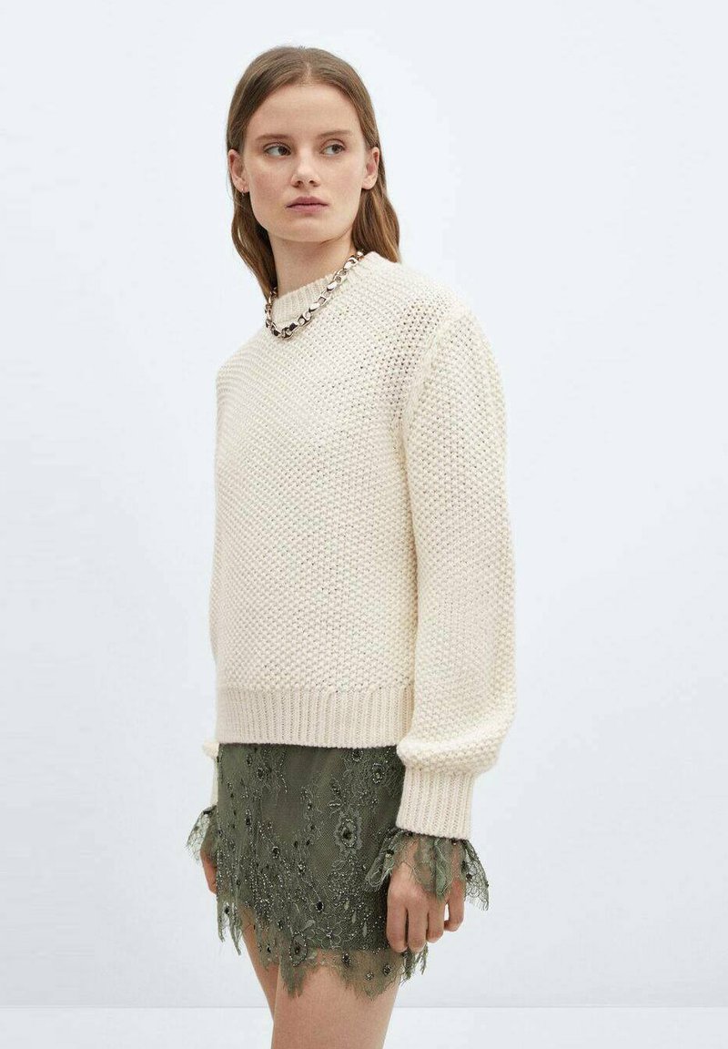 Un pull en tricot crème avec un ourlet côtelé et des manches longues, assorti à une jupe en dentelle verte foncée ornée de motifs floraux et de perles.